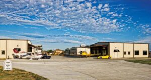 Hydraulic doors on an airplane hangar are open, it's a sunny day with blue skies, and a yellow airplane and yellow sports car are parked in front of the hangar.