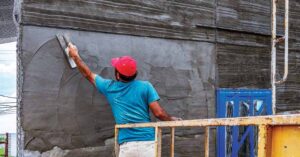 A construction worker wearing a blue t-shirt, white pants and red baseball cap is standing on scaffolding and applying materials to the exterior wall of a building. 