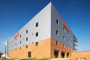 Warehouse exterior shows pewter and silver metal panels blend with brick cladding on the first floor, and windows placed throughout the four-story building.