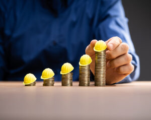 Miniature construction hats sit on top of five rows of stacked coins. From left to right, shortest stack to tallest.