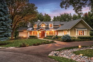 a large, white house with manicured lawn and grey roof sits beside a long driveway at dusk. 