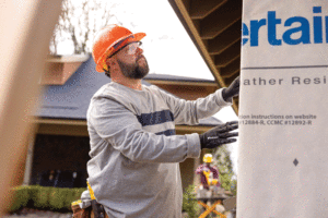 A construction worker in an orange hard hat applies white house wrap to the exterior of a house. 