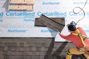 CertainTeed brand white underlayment with blue lettering is applied to a roof by a construction worker. 