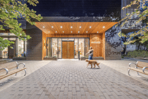 A woman walks her labrador dog on a coblestone sidewalk in front the Natiivo entrance, which features wood doors and accents. 