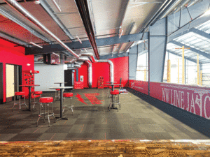 A room with high tables and chairs and grey tiled floors. The chairs have red back rests to match the red wall and red trim throughout the space. On the left are large windows letting in natural light.