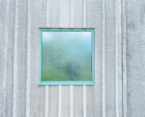 A view of a square window covered in a peelable coating. The window is trimmed in a light blue metallic and sits inside a ribbed concrete wall. 