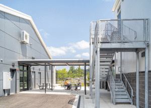 The exterior of the facility shows a metal staircase with a metal canopy, connecting to another metal building. A blue sky scattered clouds, and trees are in the background. Staff members sit at a picnic table under the canopy.