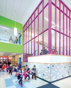 Inside an elementary school, children are waling near a stairwell  where the windows are accented with metal magenta frames. 