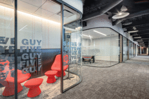 Two glass-walled offices are shown, with carpeted hallways and red, modern tables and chairs. The Five Guys logo is in white on the glass walls. 