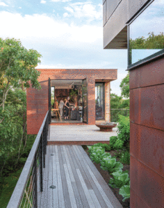 A couple is shown in their art studio. A wooden walkway leads to a wooden patio in front of the studio. The exterior is clad in weathered copper. Lush trees line both sides of the walkway.