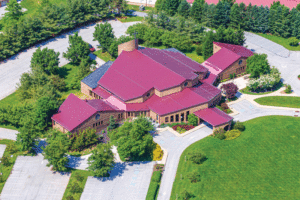 An aerial view of a mansion features a dark red metal roof and is surrounded by a large driveway and manicured lawn. 
