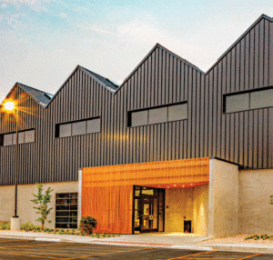 A horizontal view of a building. The lower half is concrete and the top half is brown metal panels with a metal roof that peaks at four points. 