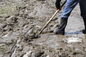 A restoration worker is shown wearing rubber boots, cleaning up a muddy area with a shovel.