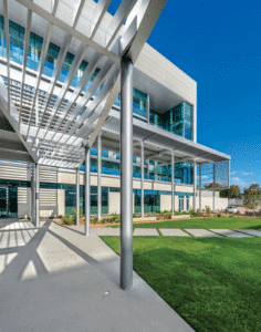 An exterior view of the white panelled building shows a metal support beam, large, blue tinted windows and a vibrant green lawn out front. 