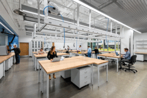 People are sitting at desks in a brightly lit lab. The ceiling is open concept and the desks are table format with wooden tops. 