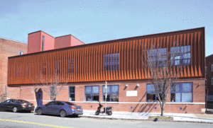 A horizontal street view of the side of the building shows orange brick on the bottom with brown/orange panels on the top half. Square windows trimmed in blue are throughout, and in front of the building is a parked car and motorcycle. 