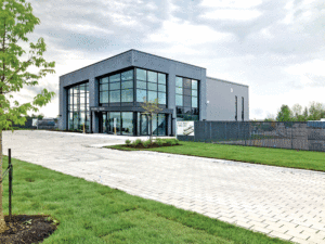 A zoomed-out angled view shows the grey panelled building with large corner windows. A cobblestone pathway leads to the building with a green lawn on either side. 