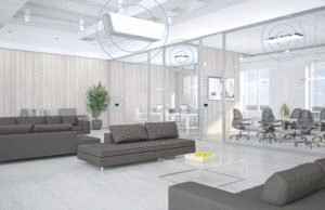 This image shows a spacious lounge area in an office building. Grey sofas sit on a white tile floor, glass office doors surround the area.