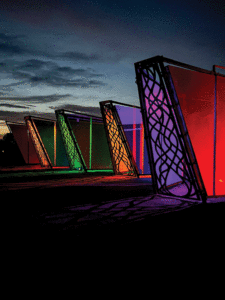 Taken at night, this outdoor shot shows four staggered metal wing shaped sculptures lit from within with colorful lights. From front to back are red, orange, green, and orange again. 