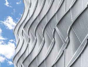 This photo shows a close-up view of wavy, perforated metal panels which make up the side of the parking garage. Behind it a bright blue sky with a few clouds. 