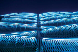 Shot from below at night, this photo shows a close-up, horizontal view of the blue LED lighting, which highlights the perforated metal on the side of the parking garage.  
