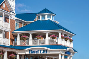 The bright blue metal trim and roofing panels compliment the white pillars surrounding the balcony. Hanging purple plants adorn the balancy and the hotel's logo, "Hotel LBI" is also showcased in bright blue against white panels. In the background is a blue sky. 