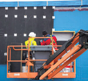 This image shows two construction workers on an orange crane applying panelling to a building.