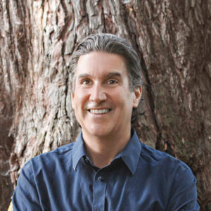 In this headshot of the author Alan Scott, he is wearing a dark blue dress shirt, leaning against a big oak tree.