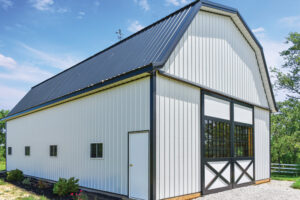 This image shows an angled view of a white panelled barn with a grey roof and dark grey trim. It is a sunny day with a blue sky with a few clouds in the background.
