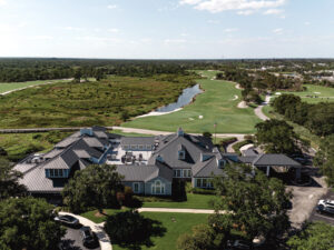 This image shows a zoomed-out, aerial view of the golf club and golf course. The clubhouse features grey metal roofing. Lush green trees surround the course and a small pond can be seen in the background. 