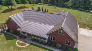 An aerial view of a large red-brick building with a brown metal roof. Surrounding the house is a large lawn with a row of trees towards the back.