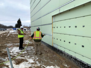 Beside a light green paneled building, two construction workers are standing on the muddy site, spraying the panels with a hose. 
