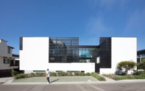 A horizontal view of a white, rectangular, one-story building with black perforated metal screening. Out front a man walks by and blue sky is in the background.