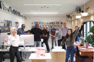 A group of people pose for the camera in an office area with desks and tables.