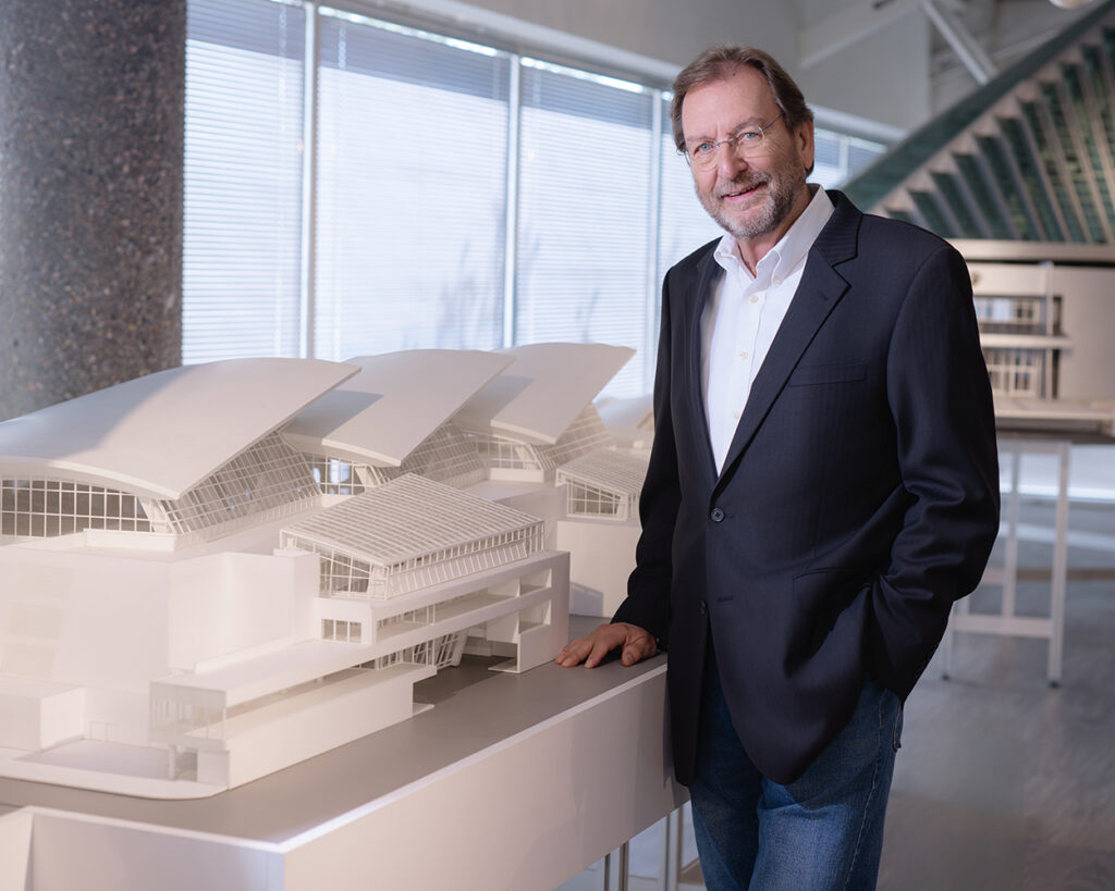 A white, bearded man stands to the right, next to a large architecture model of a structure.