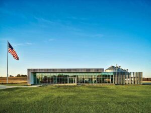 The exterior view of a one-story glass and metal building behind a green field. 