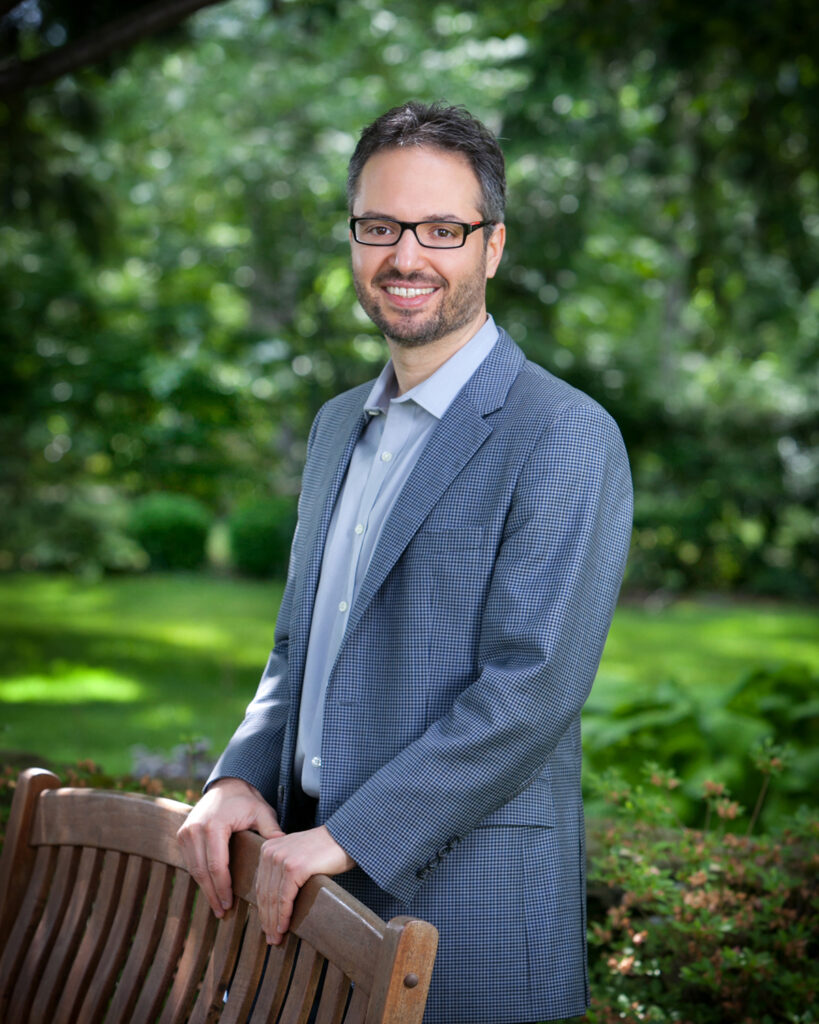 A bearded white male in a blue suit jacket leans against a park bench.
