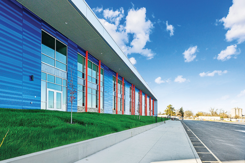 Left of a parking lot and sidewalk sits a single-story retail facility with metal cladding and glass doors.