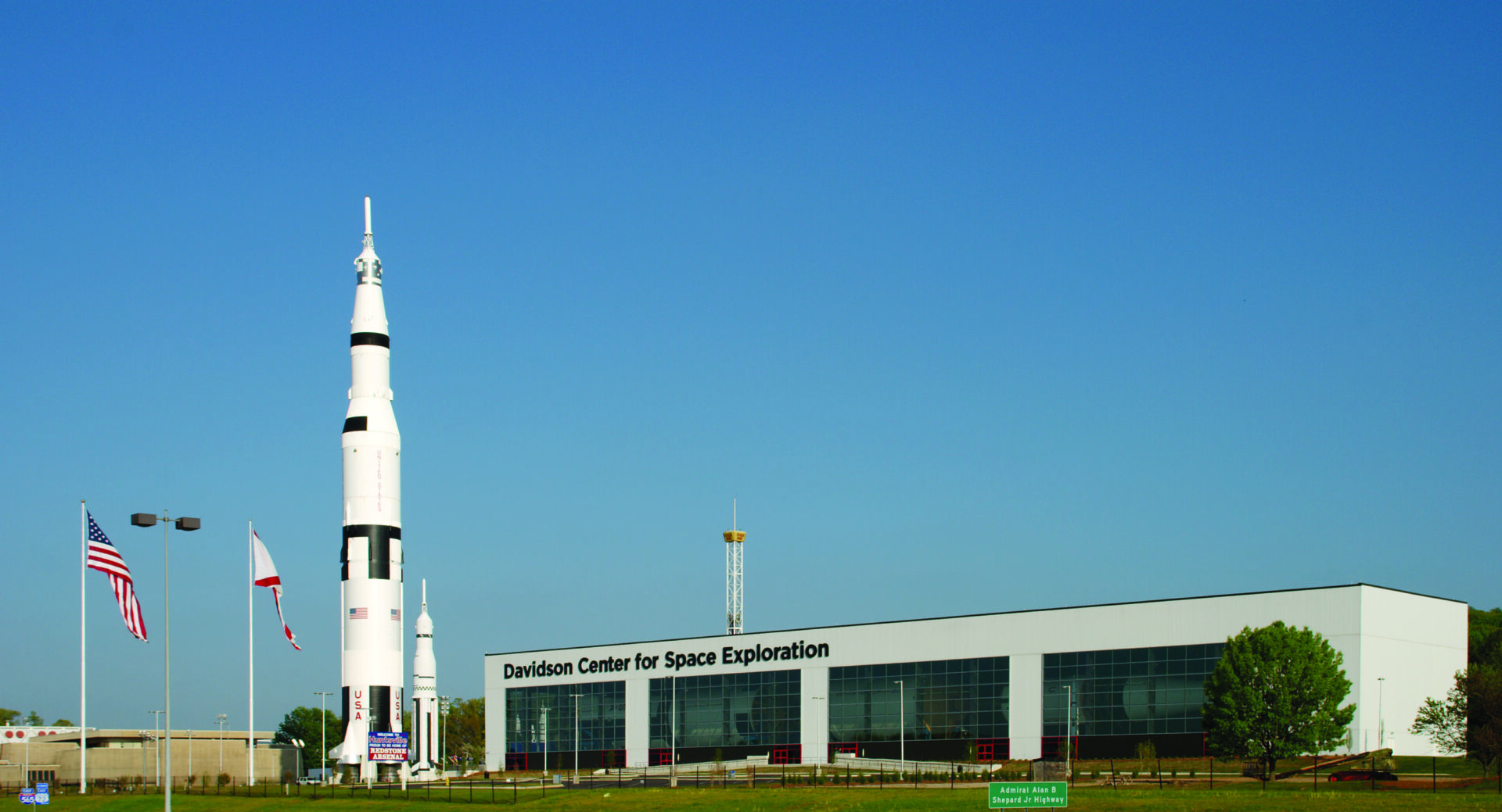 A model of the Saturn V rocket stands next to a rectangular museum and two flag poles flying the American and Alabama flags.