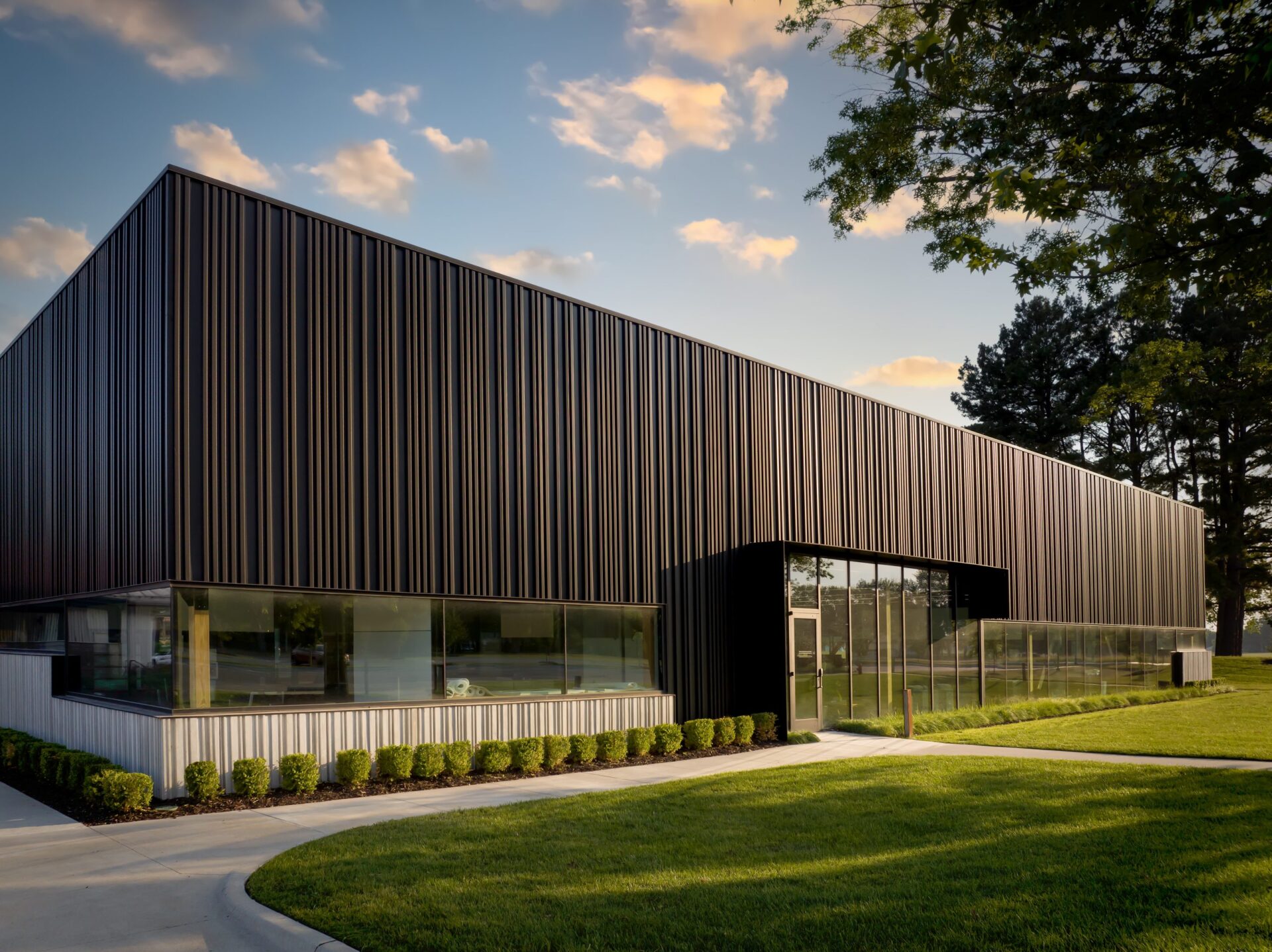 The sun sets on an office building with corrugated wall panels. In front of it is a sidewalk curving around a grass lawn.