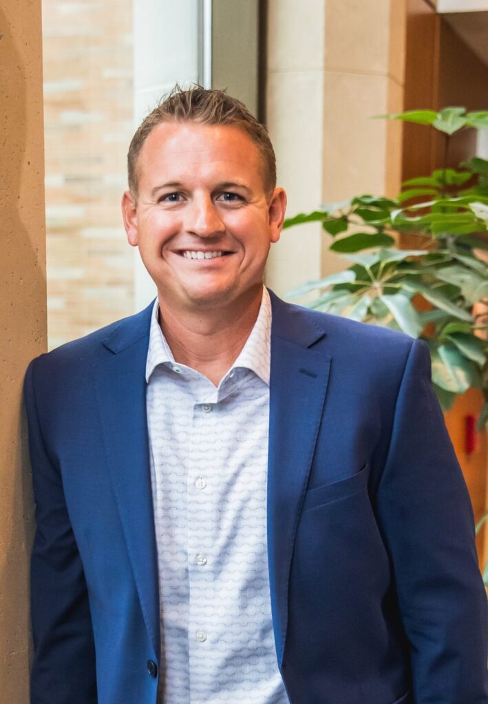 A clean-shaven man with close-cropped hair, wearing a collared shirt and open suit jacket smiles towards the camera.