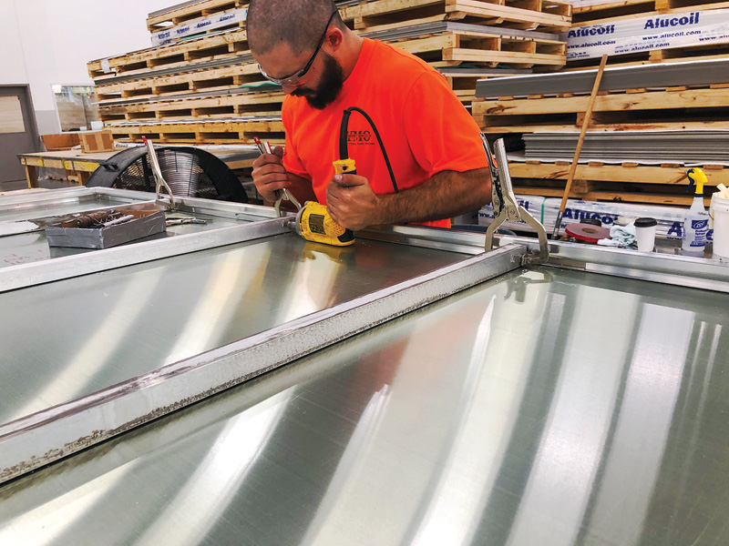 A worker wearing a t-shirt and safety glasses operates a small device to flatten a sheet of metal.