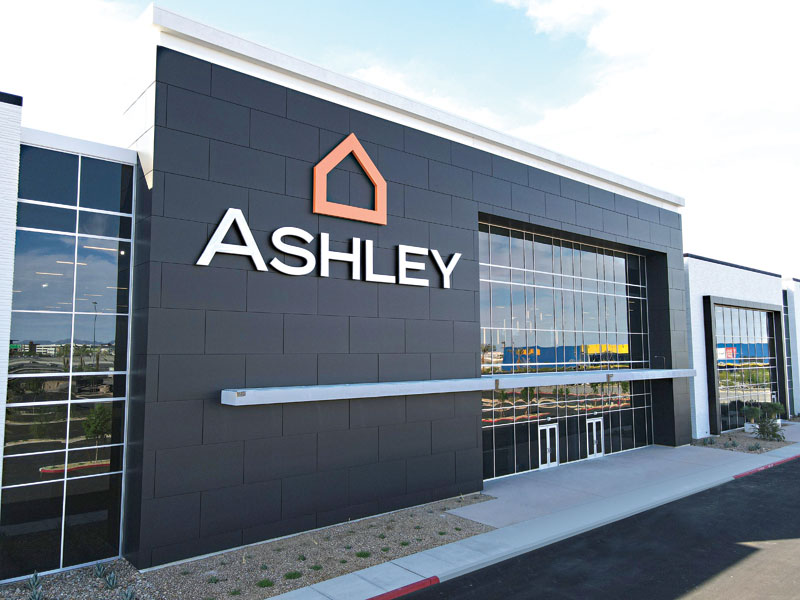 A sleek retail outlet has metal walls around a glass doorway section. Letters on the metal facade spell the company name 'Ashley' along with its logo.