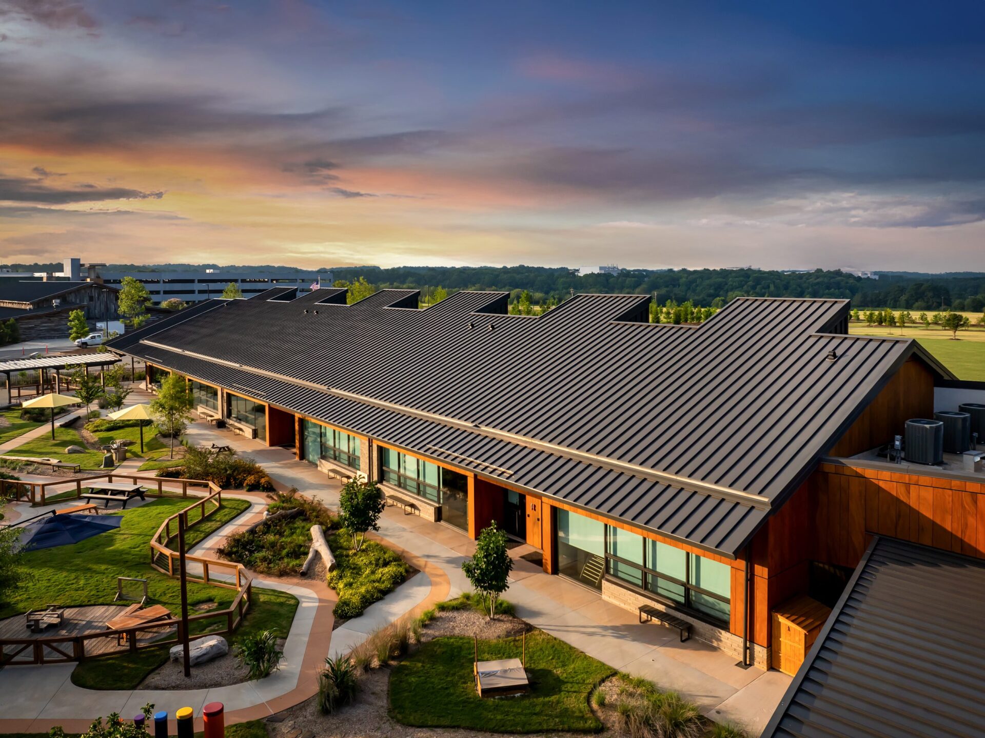 A single-story corporate building with a rustic look has a network of concrete walking paths that extend from it into a garden courtyard while the sun sets in the background.