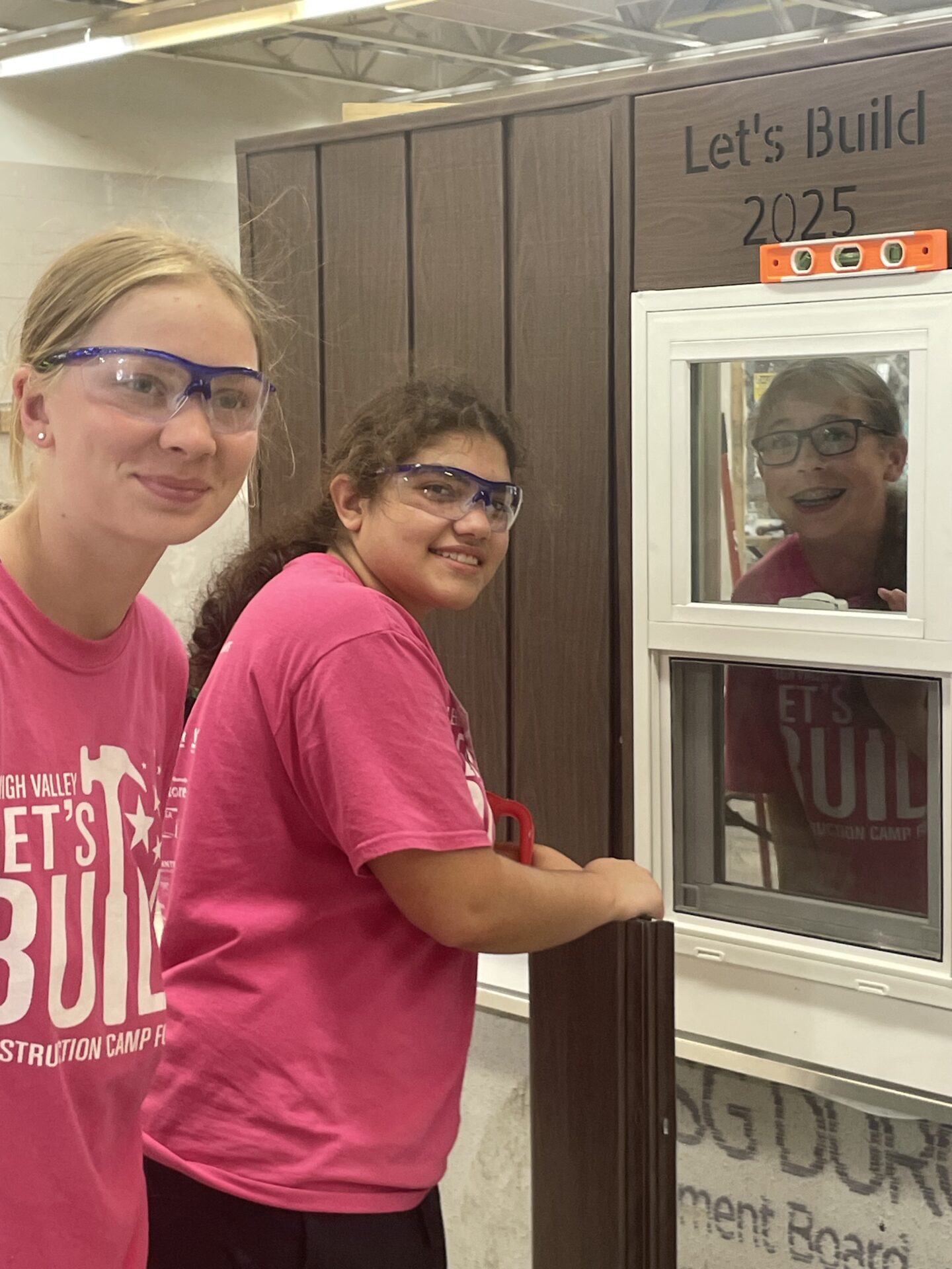 Two girl campers smile into the camera while installing metal wall panels on an educational shed. One of the female camp counselors smiles from behind a window in the shed.