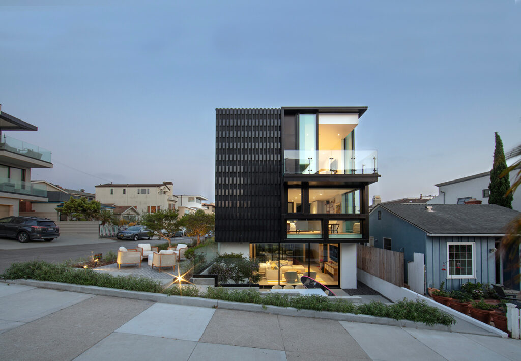 Modern three-story home with glass balconies and black metal facade, shown at dusk with illuminated interior and patio seating.