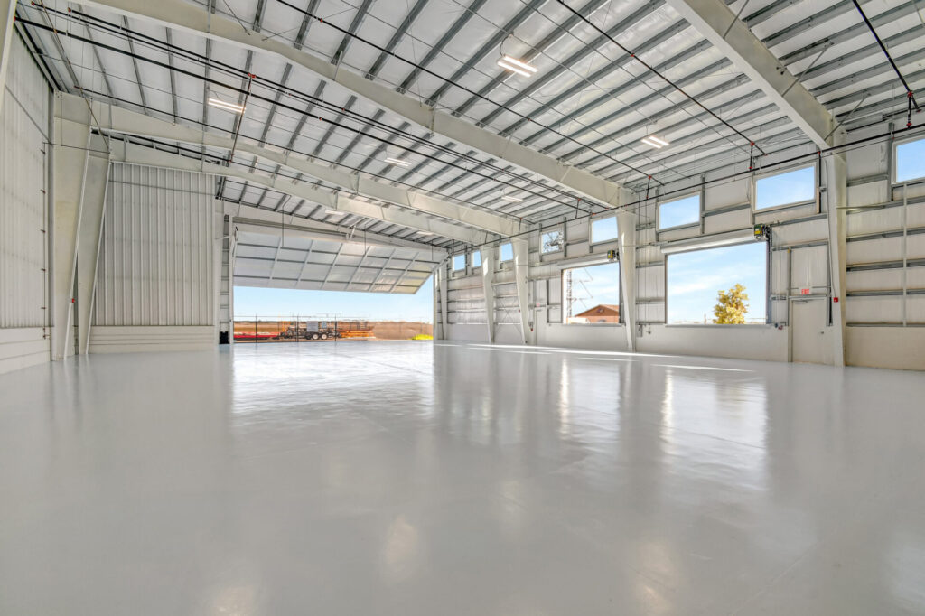 Interior of a large, empty metal hangar with polished floors, high ceilings, and a wide hydraulic door open to the outside.