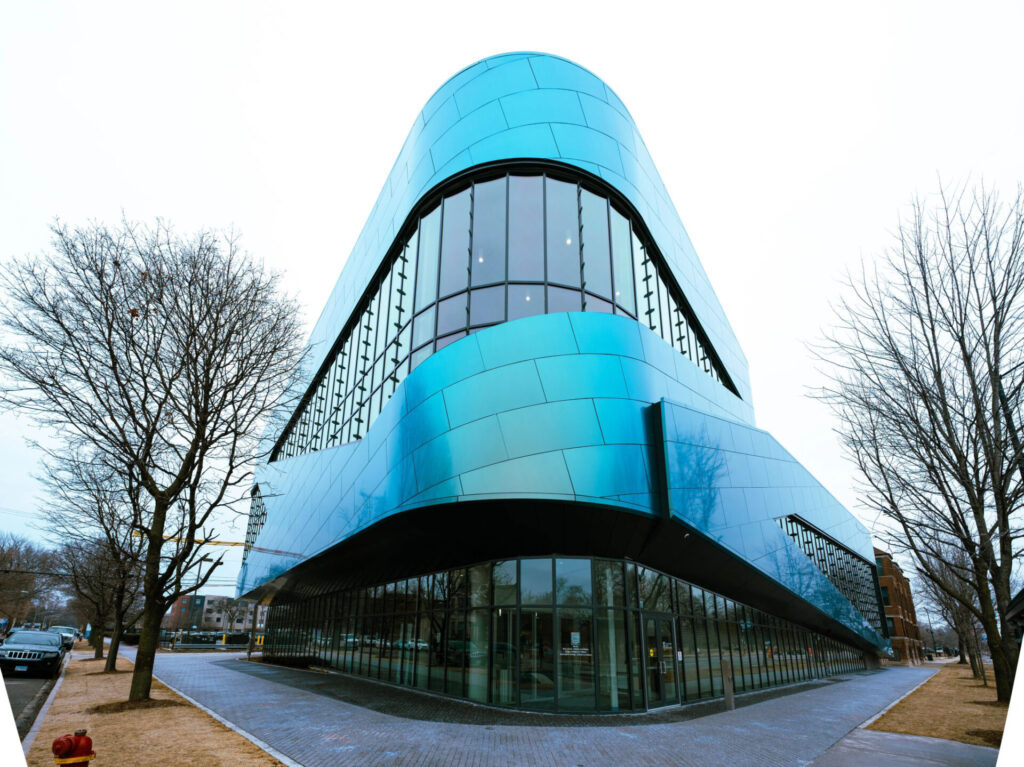 Modern building with curved blue metal panels and floor-to-ceiling glass, viewed from a low angle on a cloudy day.