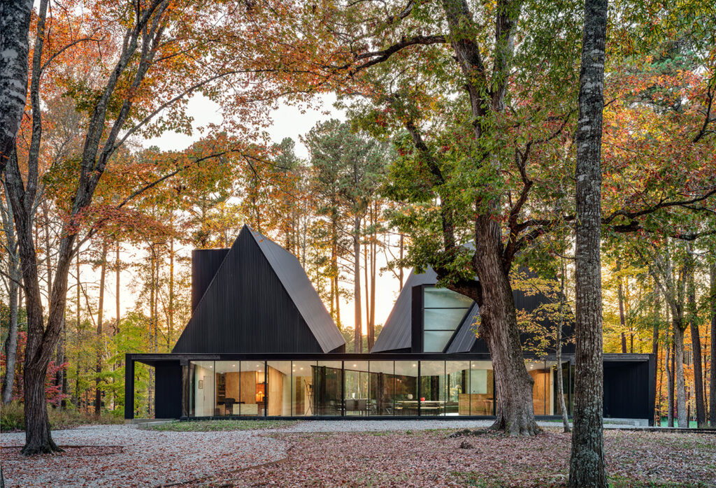 Modern black metal house with angular roofs and floor-to-ceiling glass walls, set in a wooded forest during autumn.