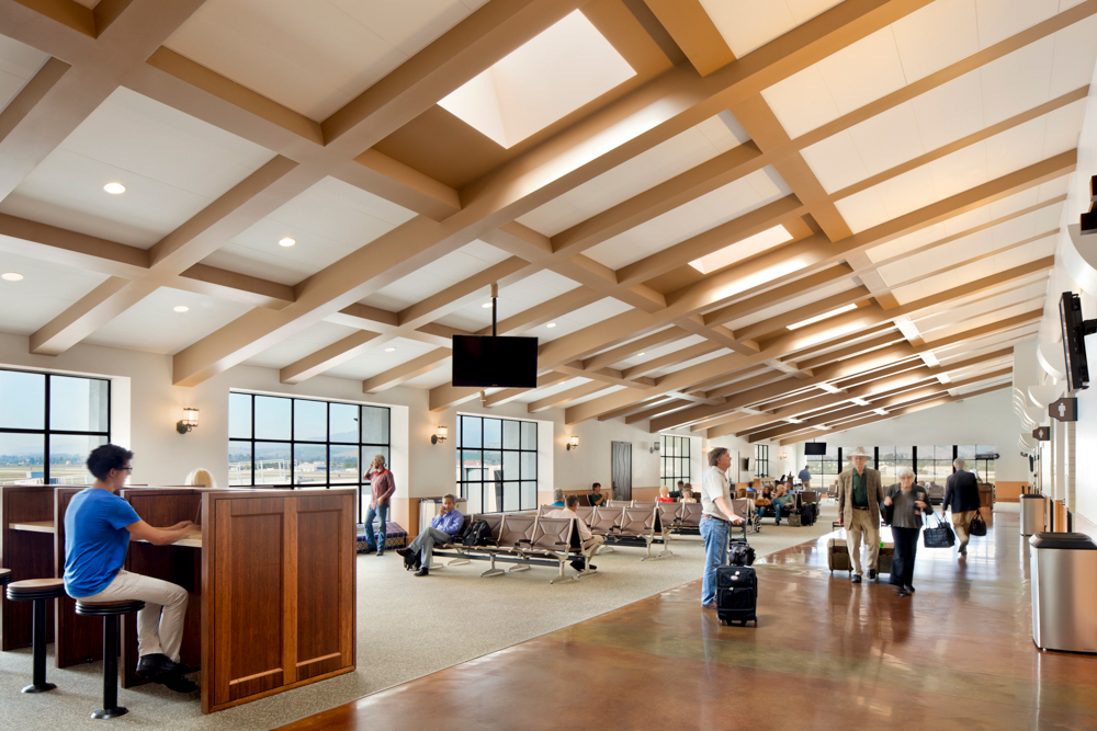 Spacious airport concourse with coffered ceiling, natural light, large steel windows, and travelers walking and seated.